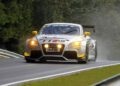 A silver and yellow Audi TT RS race car, featuring the iconic Audi five-cylinder engine, drives on a wet racetrack surrounded by trees, with headlights and fog lights on.