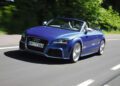 A blue Audi TT convertible, celebrating the 50th anniversary of the iconic Audi five-cylinder engine, cruises on a sunny road lined with trees in the background.
