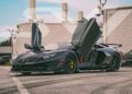 A matte black Lamborghini sports car with its scissor doors open is parked outdoors on a paved surface near modern buildings, showcasing one of the stunning exotics for sale at ABZ Motors.