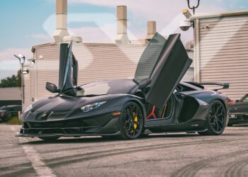 A matte black Lamborghini sports car with its scissor doors open is parked outdoors on a paved surface near modern buildings, showcasing one of the stunning exotics for sale at ABZ Motors.