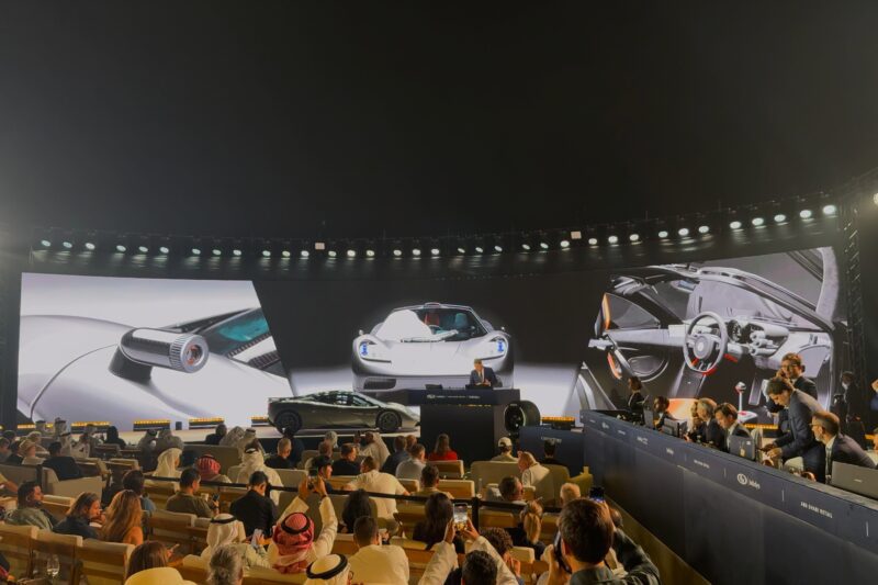 A crowd watches a car unveiling event indoors, with a sports car from Halo Cars Group displayed on stage and large screens showing close-up images of the vehicle.