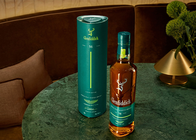 A Glenfiddich 16-year-old single malt Scotch whisky bottle and its teal cylindrical box are displayed on a green marble table beside a vase with green flowers, perfect for the 2026 duPont REGISTRY Holiday Gift Guide.