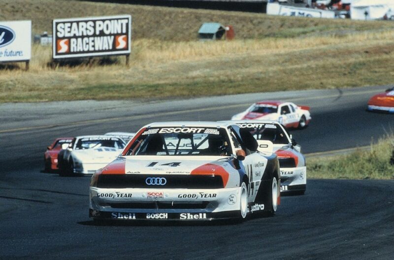 Several race cars drive on a paved track at Sears Point Raceway, with a Retro-inspired Audi RS3 leading the pack; dry grass and a track sign are visible in the background.