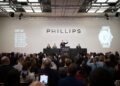 An auctioneer conducts a watch auction at Phillips, with attendees photographing the event. Currency values and an image of one of the most expensive watches are displayed on large screens during this record-breaking year.