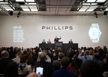 An auctioneer conducts a watch auction at Phillips, with attendees photographing the event. Currency values and an image of one of the most expensive watches are displayed on large screens during this record-breaking year.
