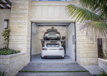 Two cars are parked in a double garage with an Autostacker car lift; a white Tesla sits below while a silver car is elevated above, maximizing garage space.