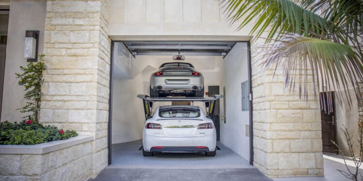 Two cars are parked in a double garage with an Autostacker car lift; a white Tesla sits below while a silver car is elevated above, maximizing garage space.