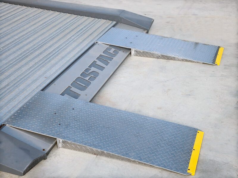 Metal loading dock ramp with two diamond-plate platforms and yellow safety edges spans a concrete floor, ideal for double garage setups or maximizing garage space, marked with the word "TOSTAG.