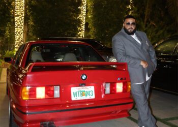 A man in a gray suit leans against a red E30 BMW M3 with a Florida license plate reading "WTBM3." Trees with string lights add to the scene, evoking the bold style of DJ Khaled.