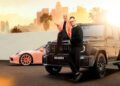 Two people pose with luxury cars in an urban setting; one sits on the hood of a black BRABUS SUV, while the other stands in front—capturing a ONE SECOND WOW moment with a pink sports car and city skyline in the background.