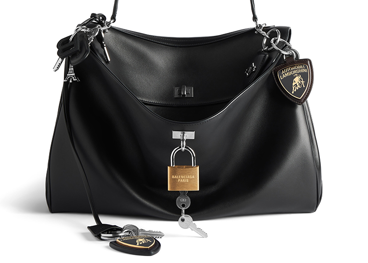 A black leather handbag with silver hardware, attached keyrings, a Balenciaga Paris padlock, and Lamborghini-branded keychain fobs—an elevated accessory featured in the 2026 duPont REGISTRY Holiday Gift Guide.