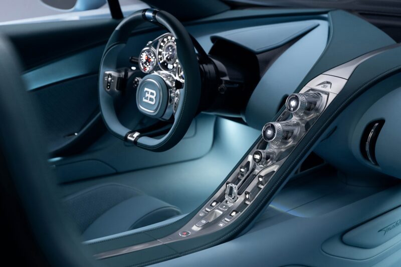Close-up view of a Bugatti Tourbillon interior showcasing the steering wheel with logo, detailed dashboard, and metallic center console with various controls—pure hypercar luxury.