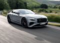 A silver Bentley coupe with a carbon fiber styling package glides along a winding rural road, surrounded by lush greenery and rolling hills in the daylight.