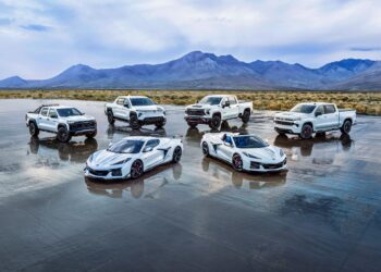 Six white vehicles, including three pickup trucks and three sports cars from the Chevy Honors America’s 250th Anniversary Ultra-Exclusive Stars & Steel Collection, are parked on a wet salt flat with mountains in the background under a cloudy sky.