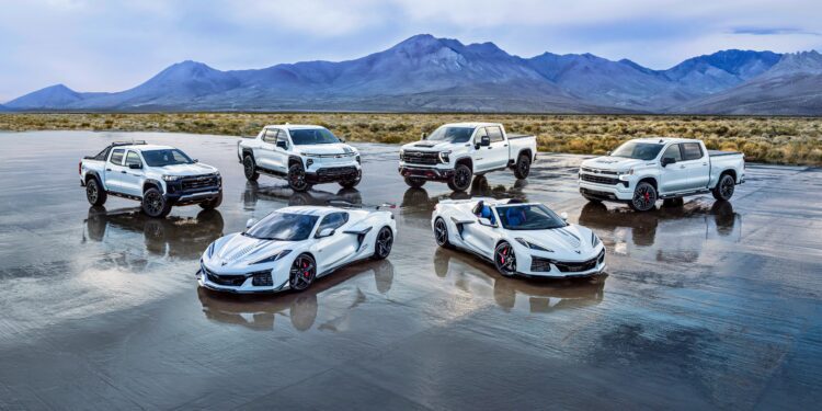 Six white vehicles, including three pickup trucks and three sports cars from the Chevy Honors America’s 250th Anniversary Ultra-Exclusive Stars & Steel Collection, are parked on a wet salt flat with mountains in the background under a cloudy sky.