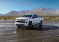 A white pickup truck from the Chevy Honors America’s 250th Anniversary With Ultra-Exclusive Stars & Steel Collection is parked on a flat, reflective surface with mountains and blue sky in the background.
