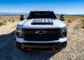 A white Chevrolet pickup from the Chevy Honors America’s 250th Anniversary With Ultra-Exclusive Stars & Steel Collection, featuring a black grille and an American flag decal, is parked in a desert landscape under a clear blue sky.