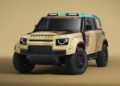 Beige and brown off-road Defender D7X-R with racing livery, rally decals, roof lights, and rugged tires, displayed in a studio setting with a plain beige background.