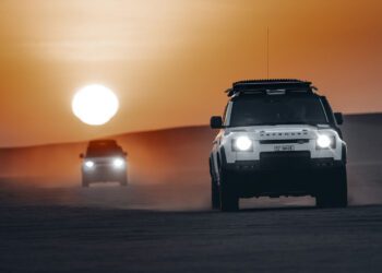 Two Defender vehicles race across sandy terrain at sunset, dust trailing behind as they follow their route planning, evoking the thrill of the Dakar Rally.