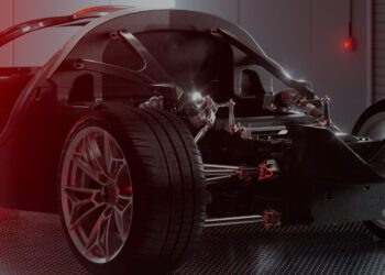 Close-up of an Apollo EVO Hypercar chassis and suspension system in a modern, well-lit workshop, highlighting the monocoque structure, visible tires, and metal frame components.