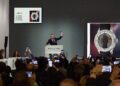 An auctioneer holds a gavel at a Phillips auction, with images and details of a wristwatch displayed on screens and attendees taking photos with phones.