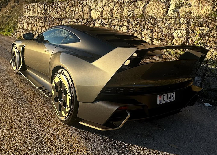 Matte black sports car with wide body kit, large rear wing, and custom wheels parked on a sunlit road beside a stone wall.
