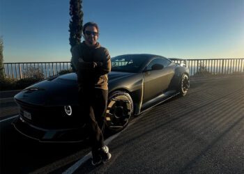 A person wearing sunglasses stands with arms crossed in front of a black sports car parked on a roadside with a metal railing and clear sky in the background.