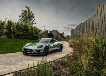 A green sports car is parked on a paved driveway beside modern fencing and landscaping under a cloudy sky.