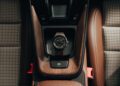 A wristwatch with a dark face and brown strap, part of a capsule collection, rests on the wooden center console of a Porsche between two brown seats with houndstooth fabric inserts.
