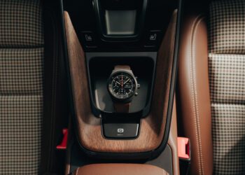 A wristwatch with a dark face and brown strap, part of a capsule collection, rests on the wooden center console of a Porsche between two brown seats with houndstooth fabric inserts.