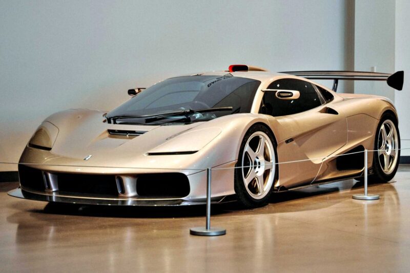A silver sports car with a low profile and large rear wing is displayed indoors behind protective barriers, evoking the spirit of the 20 Cars That Focused on Analog engagement in 2025.