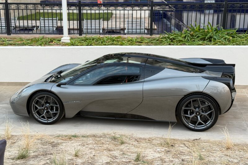 A sleek, silver sports car from Gordon Murray Automotive with black wheels and a black roof is parked on a paved surface next to a white wall and green plants.