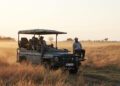 A group of people ride in an open INEOS Grenadier Game Viewer through tall, dry grass at sunset, with one person seated on the front of the vehicle.
