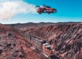 A red sports car launches off a ramp, soaring over a canyon and a moving semi-truck with trailers on a dirt road below—classic Pastrana Gymkhana Film action set in a breathtaking desert landscape.