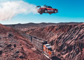 A red sports car launches off a ramp, soaring over a canyon and a moving semi-truck with trailers on a dirt road below—classic Pastrana Gymkhana Film action set in a breathtaking desert landscape.