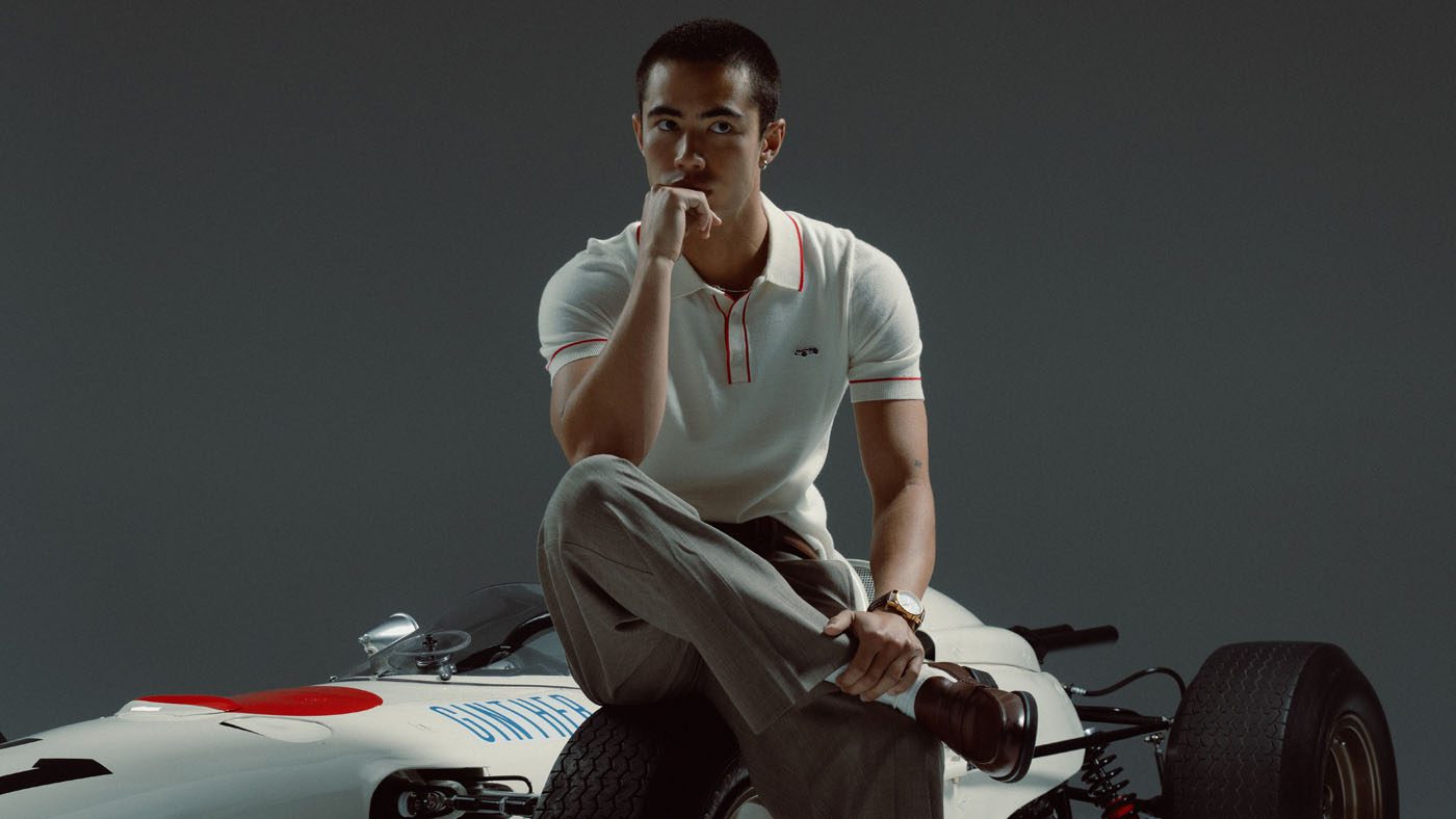 A man in a white polo shirt and grey pants sits on the front of a vintage RA272 race car, resting his chin on his hand as he looks at the camera—capturing the legacy of motorsport history.