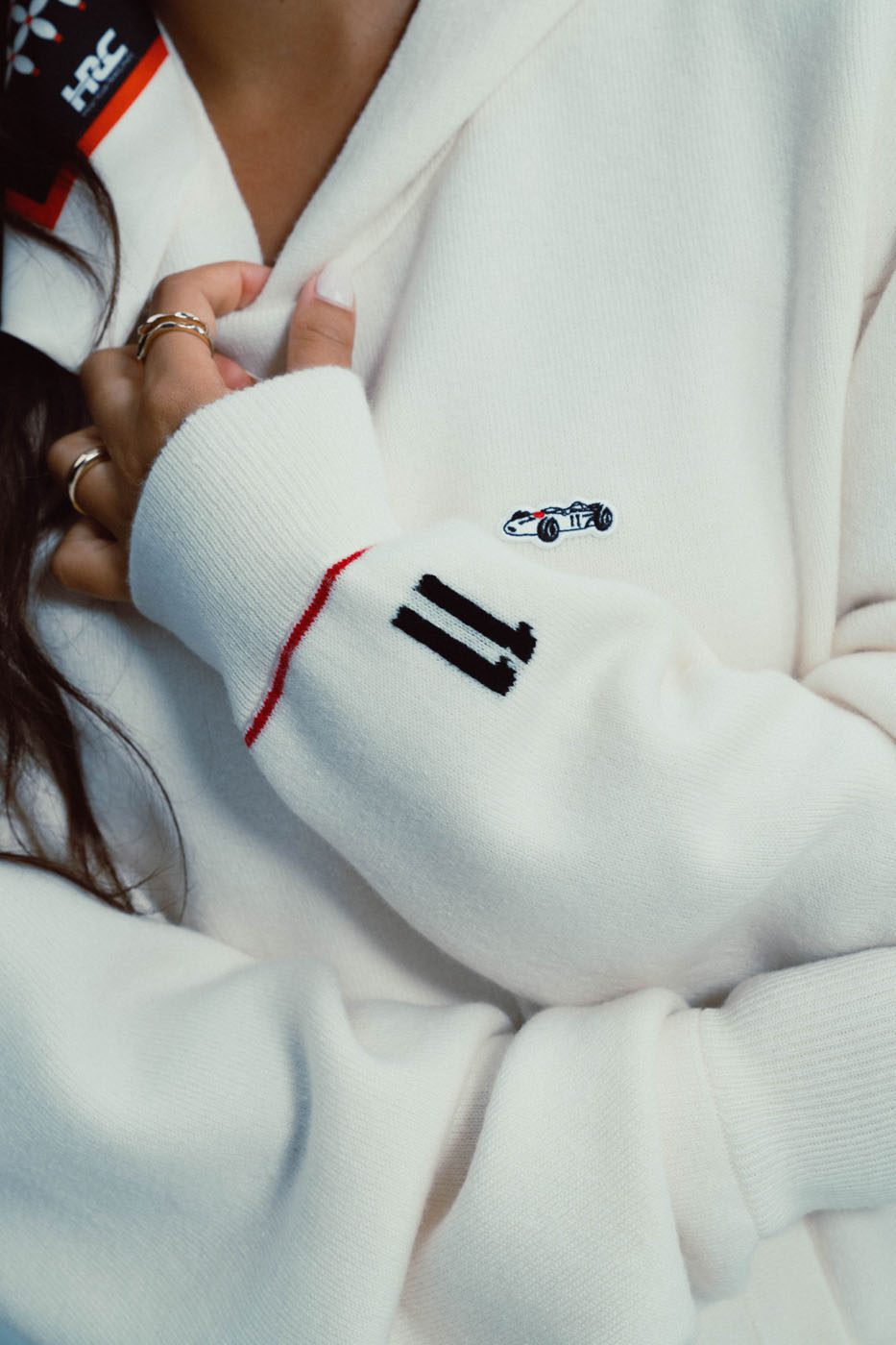 Close-up of a person wearing a white sweater with a small RA272 racecar patch, the number 11 on the sleeve, and several gold rings on their fingers—a subtle nod to racing legacy and honor.