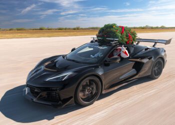 A person dressed as Santa Claus drives a 196-MPH Hennessey sports car with a Christmas tree secured on the roof, showcasing holiday spirit down an empty road.