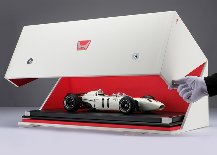 A hand in a white glove opens a white and red display case containing a detailed scale model of a vintage Honda Formula 1 race car, featured in the duPont REGISTRY 2026 Holiday Gift Guide.