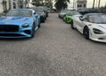 A row of luxury cars, including a blue Bentley and a white McLaren, from Dimmitt Automotive Group, parked on a palm-lined street in front of a large building during an Exotic Car Rally.