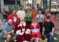 A group of people pose outdoors with two individuals dressed as Santa Claus and Mrs. Claus, along with another person dressed as an elf at the Dimmitt Automotive Group Toys for Tots event.