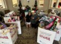 Two people kneel in front of large donation boxes filled with toys in a decorated room with Christmas trees and a chandelier during the Dimmitt Automotive Group’s Toys for Tots event.