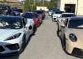 A row of sports cars, including a white Corvette and a tan Porsche, are lined up in a parking lot as part of the Exotic Car Rally hosted by Dimmitt Automotive Group, with drivers and passengers inside.
