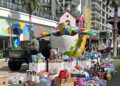 A large, colorful bird sculpture and Toys for Tots donations in boxes and piles are displayed outside a modern apartment building with palm trees.