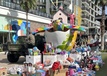 A large, colorful bird sculpture and Toys for Tots donations in boxes and piles are displayed outside a modern apartment building with palm trees.