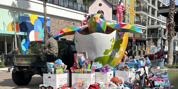 A large, colorful bird sculpture and Toys for Tots donations in boxes and piles are displayed outside a modern apartment building with palm trees.