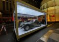 A 1937 Bugatti glistens in a glass case outdoors at night, with Rockefeller Center’s city lights and buildings illuminating the background.