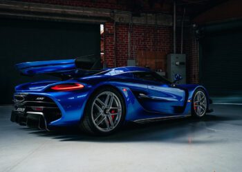 A blue Koenigsegg Jesko hypercar from O'Gara Coach is parked indoors on a concrete floor, with its rear spoiler raised and striking red brake calipers visible.