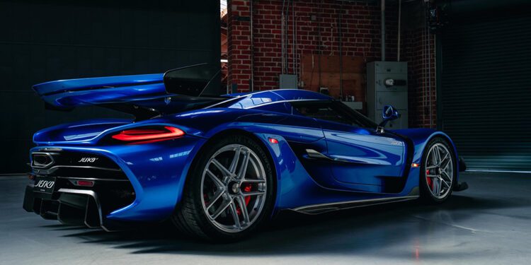 A blue Koenigsegg Jesko hypercar from O'Gara Coach is parked indoors on a concrete floor, with its rear spoiler raised and striking red brake calipers visible.