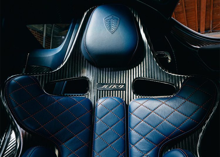 Close-up of a blue leather and carbon fiber car seat with contrast stitching, featuring the Koenigsegg Jesko hypercar logo and model name on the backrest, offered exclusively by O'Gara Coach.